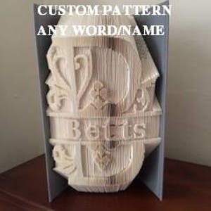 May include: A white book folded into the shape of the letter "B" with the word "Betts" written below. The book is decorated with a floral design and the words "Custom Pattern" and "Any Word Name" are written at the top.