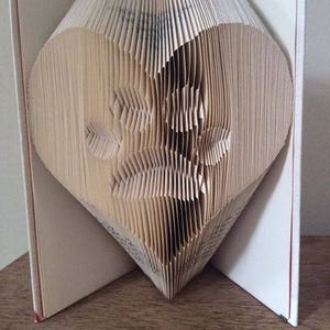 May include: A book folded into the shape of a heart. The book is made of white paper and has a cream-colored cover.