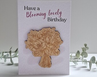 May include: A white greeting card with the text "Have a Blooming lovely Birthday" in purple script. A wooden bouquet of flowers is attached to the card. The background has a dotted pattern.