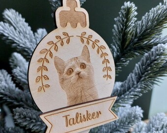 May include: A light-colored wooden Christmas ornament shaped like a bauble, featuring an engraved portrait of a cat looking upward. Above the cat is an engraved leafy branch design, and below is a banner with the name Talisker engraved in script.