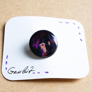 Può includere: Un bottone nero con un'immagine colorata di una persona con i capelli viola. Il bottone è su una carta bianca con la scritta "Gambit" sopra.