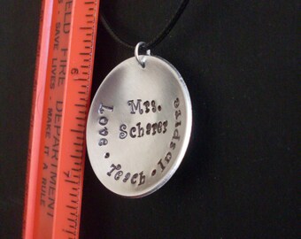 May include: A silver pendant with the words "Mrs. Scherer, Love, Teach, Inspire" stamped on it, suspended from a black cord. The pendant is circular and has a slightly textured surface. A red ruler is in the background.