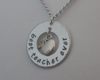 Op de afbeelding: Zilveren ketting met een ronde hanger waarop "beste leraar ooit" staat, met een klein zilveren appelbedeltje dat in het midden hangt.