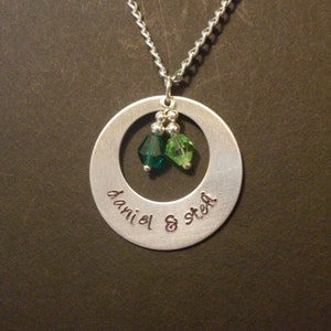 Puede incluir: Un collar de plata con un colgante redondo que tiene un círculo cortado en el centro. El colgante tiene las palabras "daniel & stef" grabadas. Dos cuentas de cristal verde cuelgan del círculo cortado.