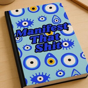 May include: A notebook with a light blue cover featuring a pattern of blue and white evil eye designs. The words "Manifest That Shit" are printed on the cover. The notebook has a black spine and is on a wooden surface.