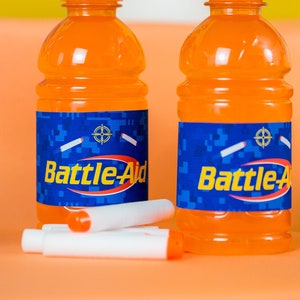 May include: Two orange plastic bottles with blue labels that say "Battle-Aid" on them. The labels have a camouflage pattern and a target symbol. The bottles are sitting on a peach-colored surface.