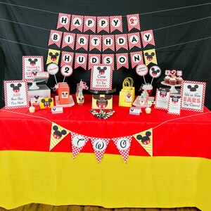 May include: A festive birthday party setup with a red and yellow tablecloth. Decorations include banners, signs, and party favors featuring a cartoon mouse character. The banner reads "Happy Birthday Hudson 2".