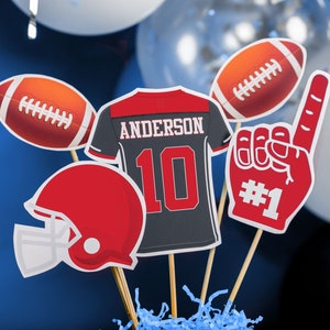 Puede incluir: Un centro de mesa con tem&aacute;tica de f&uacute;tbol con una camiseta roja y negra con el nombre "Anderson" y el n&uacute;mero "10" en ella, un casco de f&uacute;tbol rojo y blanco, un bal&oacute;n de f&uacute;tbol rojo y blanco, un dedo de espuma rojo y blanco con el n&uacute;mero "#1" en &eacute;l, y un bal&oacute;n de f&uacute;tbol rojo y blanco.