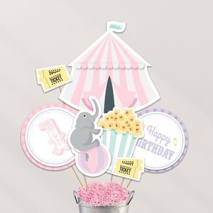 Könnte beinhalten: Ein Party-Dekorationsset zum Thema Zirkus. Enthält ein rosa-weiß gestreiftes Zirkuszelt, einen grauen Elefanten, der auf einem Ball balanciert, Popcorn, Tickets und ein "Happy Birthday"-Schild. Die Dekorationen befinden sich in einem silbernen Eimer mit rosa Papier.