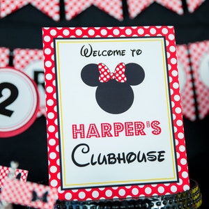以下が含まれることがあります： 赤と白のドット柄の縁取りと「Welcome to Harper's Clubhouse」の文字が入ったパーティーサイン。ミッキーマウスの頭の黒いシルエットと赤い水玉模様のリボンが特徴です。「THDA」という文字が入ったバナーが背景にあります。