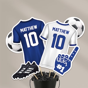 May include: Soccer-themed decorations featuring a blue jersey and a white jersey, both with the name "MATTHEW" and the number "10". Includes a pair of black soccer cleats, a foam finger, and soccer balls. All items are arranged in a black bucket.