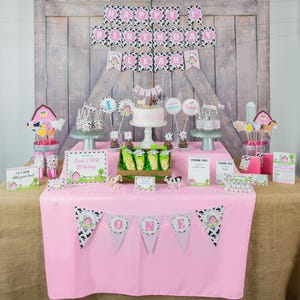 May include: A pink table with a farm-themed birthday party setup. The table is decorated with a pink and black cow print banner, a cake with a farm animal topper, and various farm animal decorations. The table also has a sign that says "Leah's First Birthday".