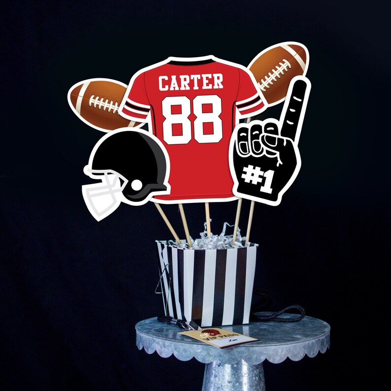 May include: A football-themed centerpiece with a red jersey with the name "CARTER" and the number "88" on it, a black football helmet, a brown football, a black foam finger with the number "#1" on it, and a black and white striped container.