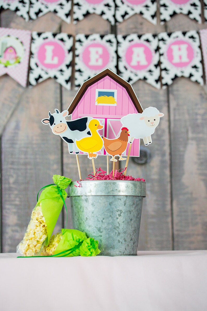 Puede incluir: Un granero rosa con un pato amarillo, una vaca blanco y negro, una oveja blanca y una gallina roja. Los animales est&aacute;n en palos y est&aacute;n en un cubo de metal con papel picado rosa y blanco. Hay bolsas de palomitas de ma&iacute;z verdes y blancas en la mesa.