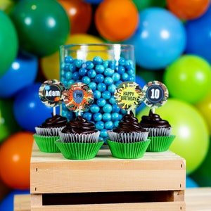 May include: Four chocolate cupcakes with green cupcake liners are on a wooden crate. The cupcakes are decorated with round toppers that say "Adam", "Happy Birthday", "10", and a camouflage pattern with a paintball mask. A glass jar filled with blue candy is behind the cupcakes.