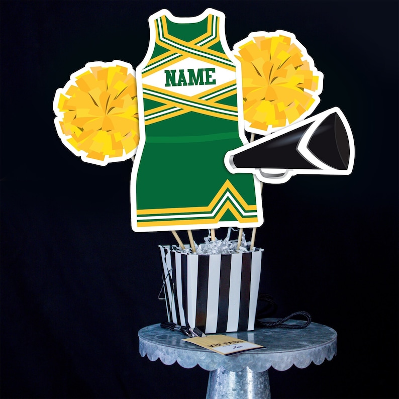 May include: Green and gold cheerleader outfit with a white name tag that says "NAME" on it. The outfit is part of a centrepiece with two yellow pom-poms and a black megaphone.