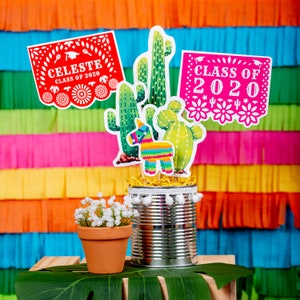 Op de afbeelding: Een feestelijk afstudeermiddelpunt met een zilveren blik met witte pompoenen, een kleine terracotta pot met witte bloemen en papieren uitsnijdingen van cactussen, een pi&ntilde;ata en banners met de tekst "Celeste Class of 2020" en "Class of 2020".