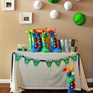 May include: A white table with a green and white checkered tablecloth and a blue ribbon banner that says "PRO SHOP". There are green and white golf-themed decorations on the wall behind the table. There are also several toy golf clubs and bags on the table.