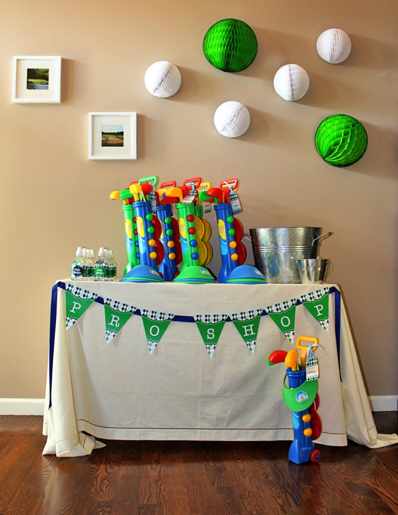 May include: A white table with a green and white checkered tablecloth and a blue ribbon banner that says "PRO SHOP". There are green and white golf-themed decorations on the wall behind the table. There are also several toy golf clubs and bags on the table.