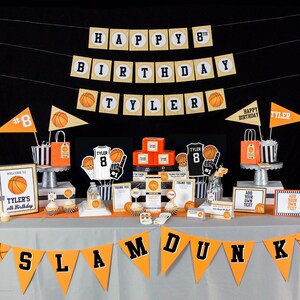 May include: A basketball-themed birthday party table setting with orange and black decorations. The table is covered in a gray tablecloth with a banner that reads "SLAM DUNK". There are several decorations, including a basketball-shaped cake, a basketball-shaped pi&ntilde;ata, and a basketball-shaped balloon. The table is also decorated with orange and black streamers and balloons.