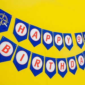 May include: A blue and red banner with the words "Happy 9th Birthday" in white circles. The banner is strung on a white string and is hanging against a yellow background.