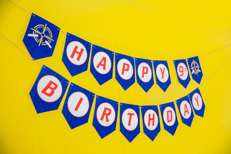 May include: A blue and red banner with the words "Happy 9th Birthday" in white circles. The banner is strung on a white string and is hanging against a yellow background.