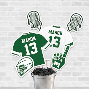 Puede incluir: Decoraciones de fiesta de lacrosse en verde y blanco. Las decoraciones incluyen un palo de lacrosse, un casco de lacrosse, una camiseta con el nombre "MASON" y el número "13", y un dedo de espuma con el número "#1".