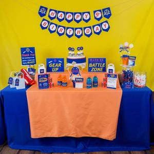 May include: A birthday party table setting with a blue and orange tablecloth. The table is decorated with a 'Happy 9th Birthday' banner, a 'Gear Up' sign, a 'Warning Battle Zone' sign, and a cake with blue and white frosting. There are also several other decorations, including cupcakes, candy, and drinks.