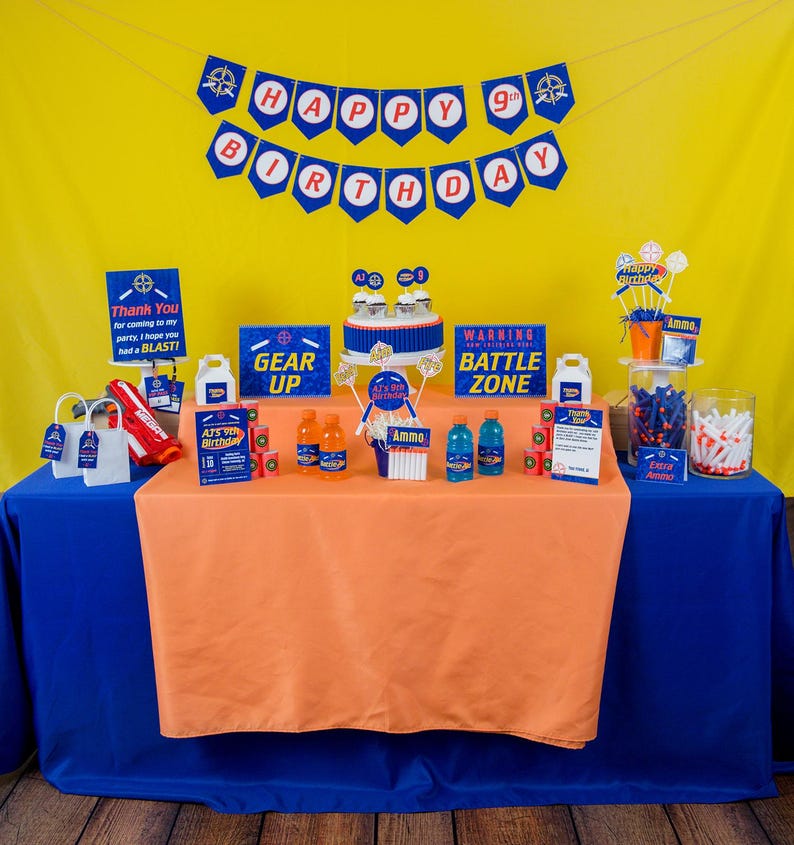 May include: A birthday party table setting with a blue and orange tablecloth. The table is decorated with a 'Happy 9th Birthday' banner, a 'Gear Up' sign, a 'Warning Battle Zone' sign, and a cake with blue and white frosting. There are also several other decorations, including cupcakes, candy, and drinks.