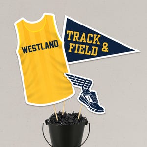 May include: A black bucket filled with black paper shreds holding three decorative cutouts: a yellow sleeveless jersey with the text WESTLAND, a dark blue triangular pennant with the text TRACK & FIELD &, and a dark blue winged running shoe.