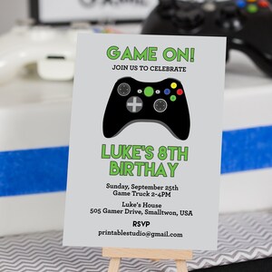 以下が含まれることがあります： 小さな木製イーゼルに置かれた灰色の招待状。カードには「GAME ON！」と「LUKE'S 8TH BIRTHDAY」と書かれており、黒いゲームコントローラーのグラフィックが付いています。招待状にはイベントの詳細とRSVPのメールアドレスが含まれています。