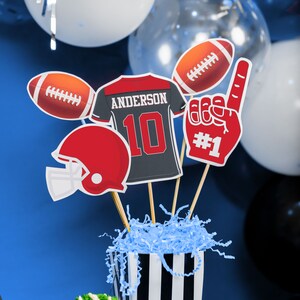 Puede incluir: Una pieza central de fiesta con tem&aacute;tica de f&uacute;tbol con una caja a rayas blancas y negras llena de papel de seda azul. La pieza central incluye una camiseta de f&uacute;tbol con el nombre "Anderson" y el n&uacute;mero "10", un bal&oacute;n de f&uacute;tbol, un casco de f&uacute;tbol y una mano de espuma roja con el n&uacute;mero "#1".