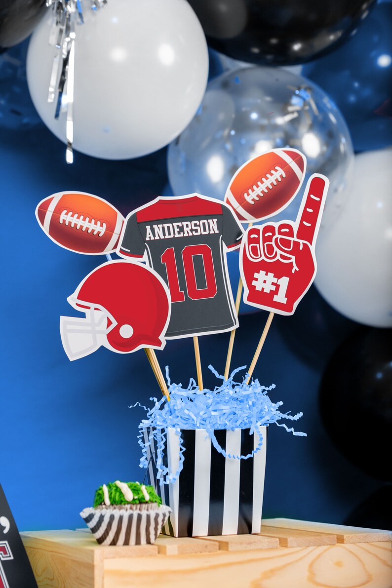 Puede incluir: Una pieza central de fiesta con tem&aacute;tica de f&uacute;tbol con una caja a rayas blancas y negras llena de papel de seda azul. La pieza central incluye una camiseta de f&uacute;tbol con el nombre "Anderson" y el n&uacute;mero "10", un bal&oacute;n de f&uacute;tbol, un casco de f&uacute;tbol y una mano de espuma roja con el n&uacute;mero "#1".