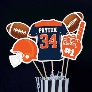 May include: A football-themed centerpiece with a blue and orange jersey, a football helmet, two footballs, and a foam hand with the number "1" on it. The jersey has the name "PAYTON" and the number "34" on it.