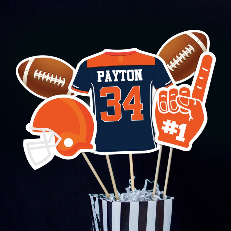 May include: A football-themed centerpiece with a blue and orange jersey, a football helmet, two footballs, and a foam hand with the number "1" on it. The jersey has the name "PAYTON" and the number "34" on it.
