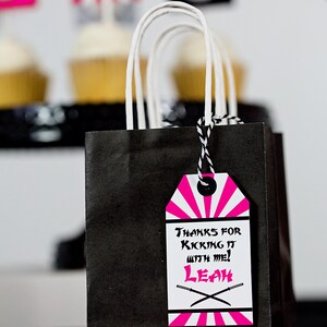 May include: Black and pink party favor bag with a tag that says "Thanks for kicking it with me! Leah" with a black and white striped design and two crossed swords.