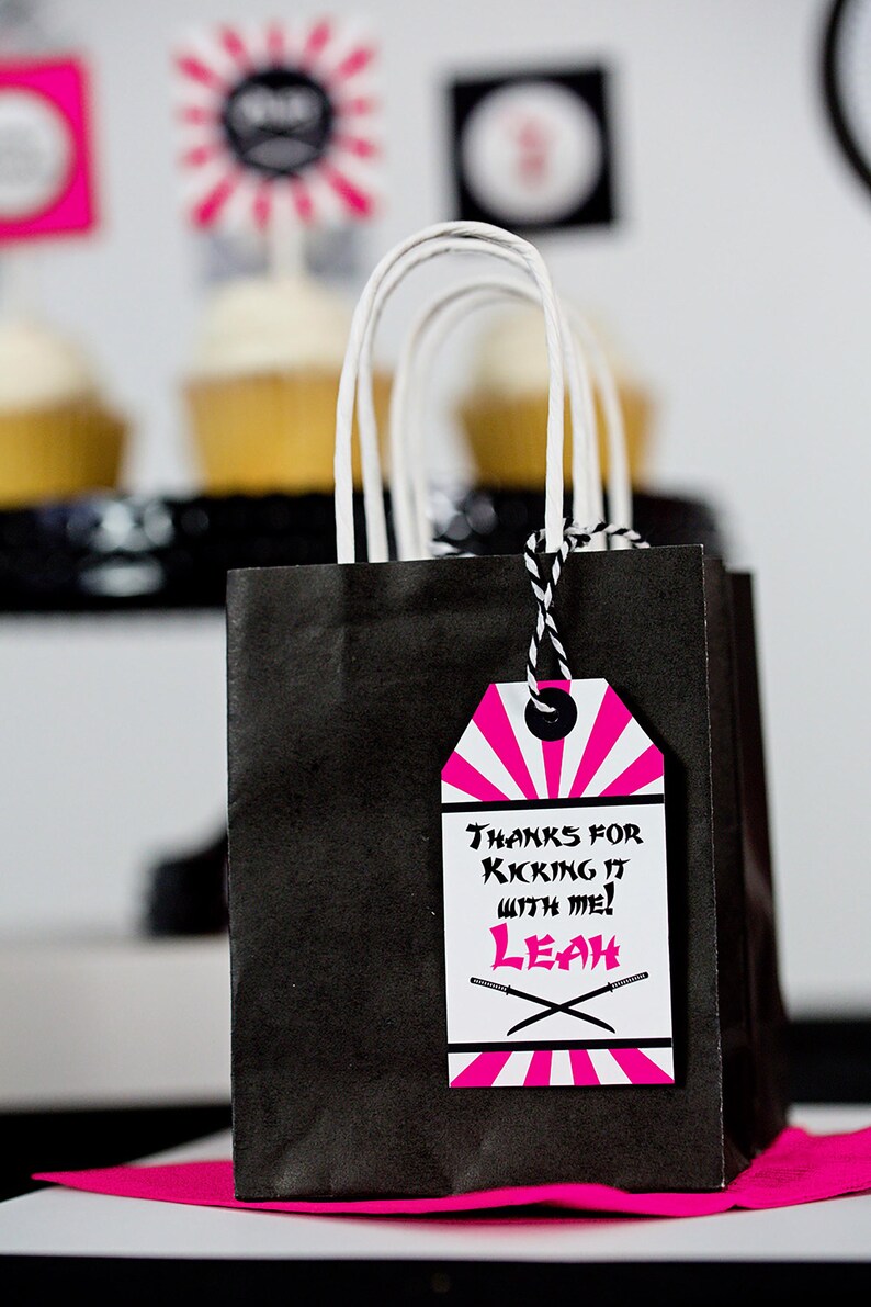 May include: Black and pink party favor bag with a tag that says "Thanks for kicking it with me! Leah" with a black and white striped design and two crossed swords.