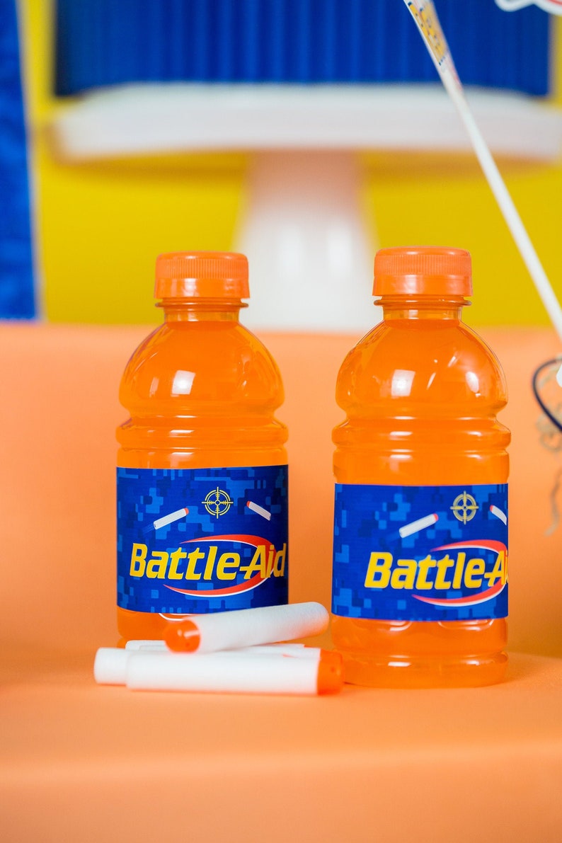 May include: Two orange plastic bottles with blue labels that say "Battle-Aid" on them. The labels have a camouflage pattern and a target symbol. The bottles are sitting on a peach-colored surface.