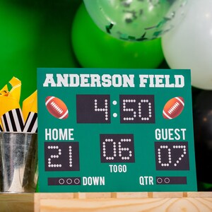 Puede incluir: Un marcador de f&uacute;tbol verde y negro con el texto "Anderson Field" y el marcador "Home 21, Guest 07". El marcador tambi&eacute;n muestra la hora "4:50", el down y el cuarto. El marcador est&aacute; sentado sobre una superficie de madera.