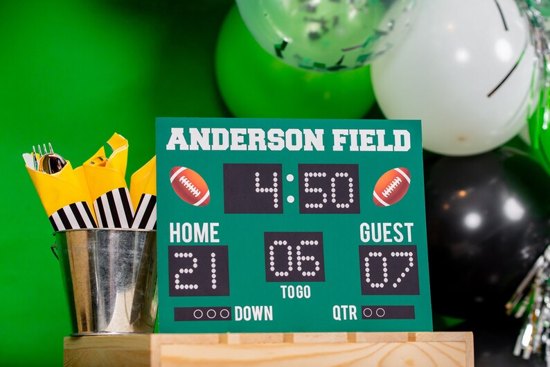 Puede incluir: Un marcador de f&uacute;tbol verde y negro con el texto "Anderson Field" y el marcador "Home 21, Guest 07". El marcador tambi&eacute;n muestra la hora "4:50", el down y el cuarto. El marcador est&aacute; sentado sobre una superficie de madera.