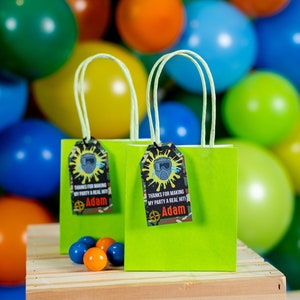 May include: Two green gift bags with lime green handles and personalized tags that say "Thanks for making my party a real hit! Adam". The bags are sitting on a wooden surface in front of a wall of colorful balloons.