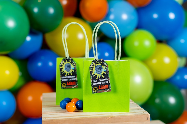 May include: Two green gift bags with lime green handles and personalized tags that say "Thanks for making my party a real hit! Adam". The bags are sitting on a wooden surface in front of a wall of colorful balloons.