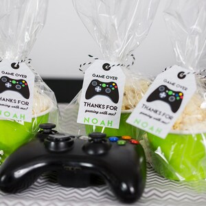 May include: Three green cups filled with popcorn and tied with clear cellophane bags. Each bag has a white tag with a black video game controller graphic and the text "GAME OVER" and "THANKS FOR gaming with me! NOAH". A black video game controller is in the foreground.