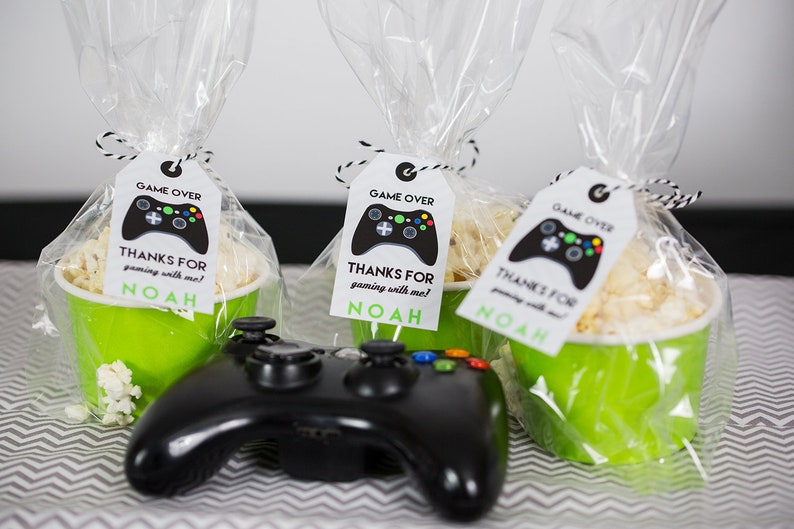 May include: Three green cups filled with popcorn and tied with clear cellophane bags. Each bag has a white tag with a black video game controller graphic and the text "GAME OVER" and "THANKS FOR gaming with me! NOAH". A black video game controller is in the foreground.