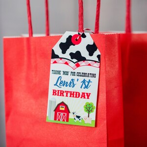 May include: Red gift bags with a cow print tag that says "Thank Moo for celebrating Lenis' 1st Birthday". The tag has a red and white checked border and a farm scene with a barn, a cow, and a pig.