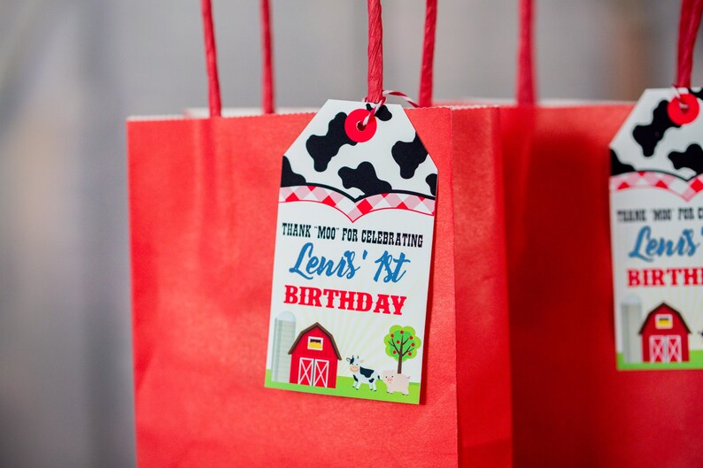 May include: Red gift bags with a cow print tag that says "Thank Moo for celebrating Lenis' 1st Birthday". The tag has a red and white checked border and a farm scene with a barn, a cow, and a pig.
