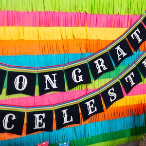 Op de afbeelding: Een zwart-witte banner met de woorden "CONGRATS CELESTE" hangt tegen een kleurrijke achtergrond van groene, roze, gele, oranje en blauwe cr&ecirc;pepapier slingers.