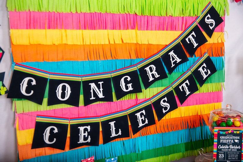 Op de afbeelding: Een zwart-witte banner met de woorden "CONGRATS CELESTE" hangt tegen een kleurrijke achtergrond van groene, roze, gele, oranje en blauwe cr&ecirc;pepapier slingers.