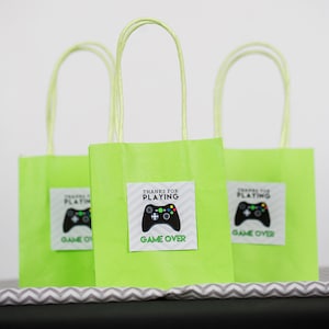 May include: Three green paper gift bags with white handles. Each bag has a sticker with a black and green video game controller and the text "Thanks for Playing Game Over".