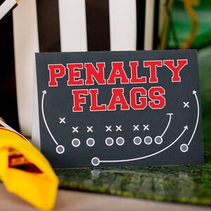 May include: A black sign with white text that reads "PENALTY FLAGS" in red letters. The sign has a football field diagram with white lines and circles.